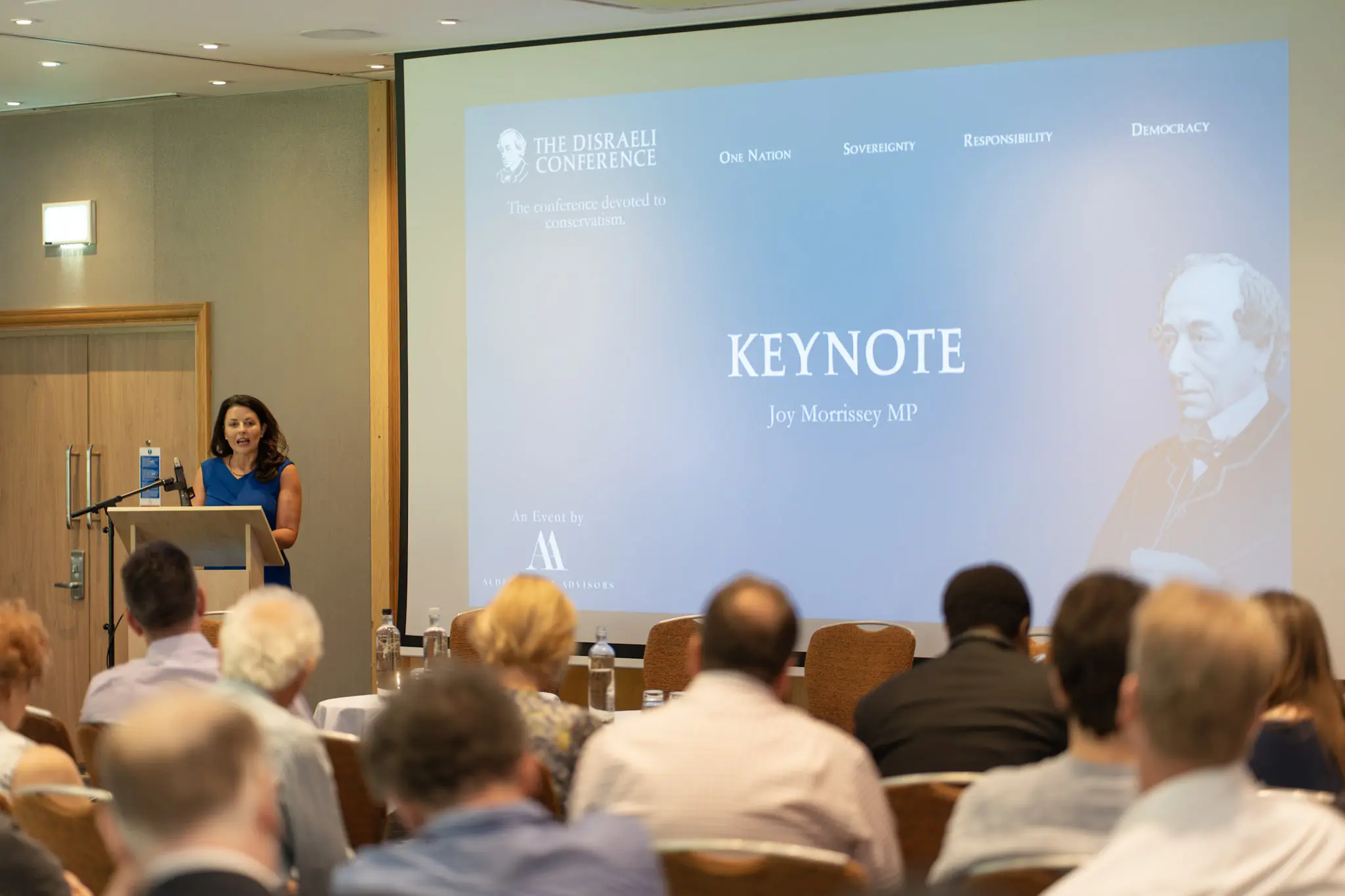 A keynote speaker at the lectern at the Disraeli Conference, beside a screen reading Keynote, Joy Morrissey MP